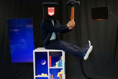 Homme assis sur une étagère qui a un masque en forme de cube et qui tient un marteau - Agrandir l'image 3 sur 3, fenêtre modale