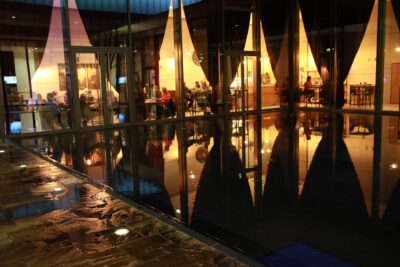 Le bistrot vu de l'extérieur, le sol est mouillé grâce à l'architecture et le bistrot est en arrière plan derrière une baie vitrée - Agrandir l'image 4 sur 7, fenêtre modale